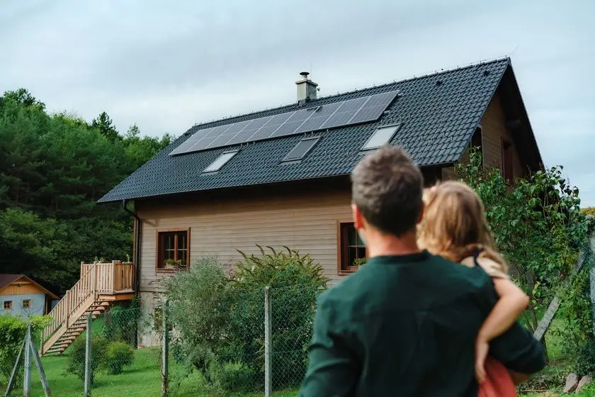Rear view of dad holding her little girl in arms and looking at their house with solar panels