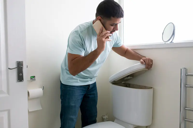 Man standing in his bathroom looking at his toilet, he is on the phone trying to call a plumber.