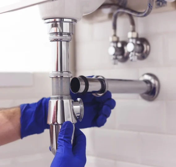 Plumber in blue gloves repairing shiny chrome pipes under a sink, illustrating The Drain Fighter's drain cleaning services.