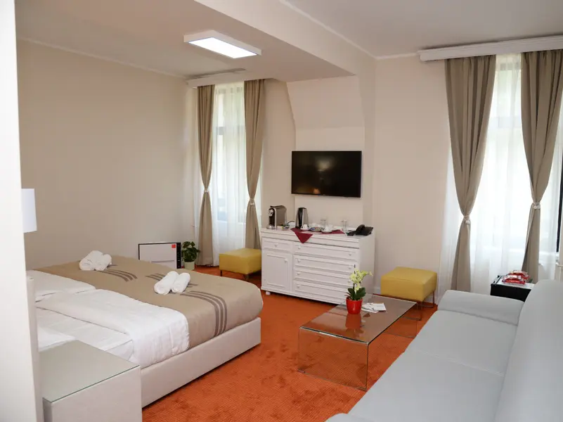 Soba u Zepter Hotel Vrnjačka Banja sa bračnim krevetom, narandžastim tepihom, belom komodom, TV-om, žutim tabureima i belim kaučem