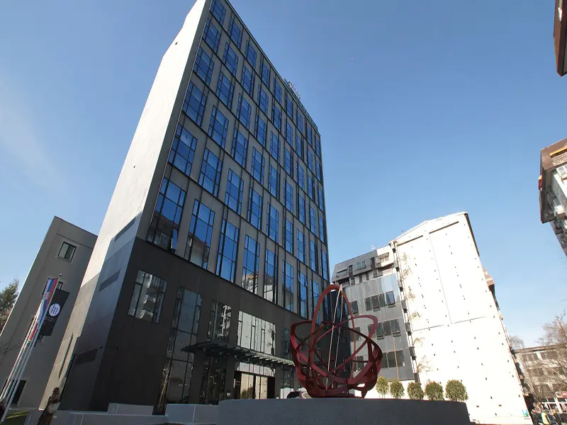 Eksterijer Hotel Mona Plaza sa modernom staklenom fasadom u tamnim tonovima i crvenom apstraktnom skulpturom ispred ulaza