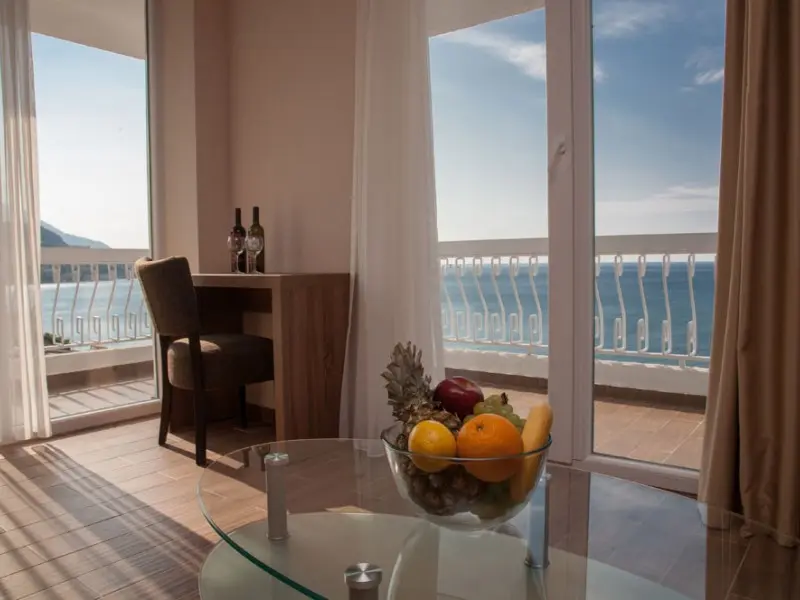 Dnevna soba apartmana u Sato Hotel Conference & Spa Resort hotelu sa voćem na staklenom stolu, vinom i panoramskim pogledom na more