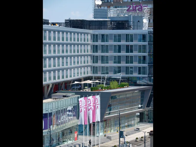 Fasada Hotel Zira Beograd danju sa modernom staklenom arhitekturom, natpisom Zira na krovu, ružičastim zastavama i zelenom terasom na krovu