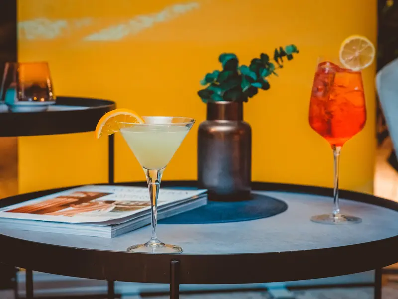Dva koktela na okruglom stočiću u baru Hotel voco Podgorica sa martini čašom, Aperol Spritzom, vazom sa eukaliptusom i žutim zidom u pozadini