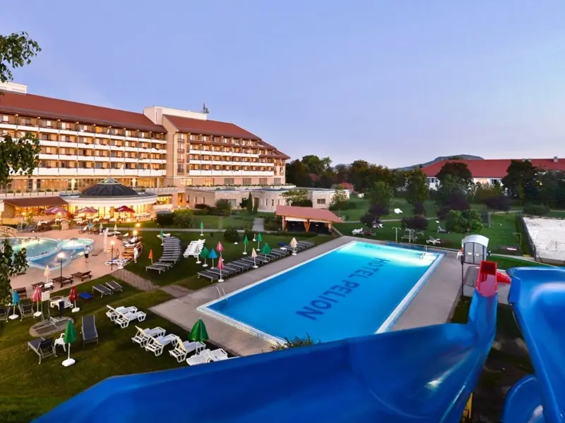 Spoljašnji pogled na Hunguest Hotel Pelion Superior sa bazenom, plavim toboganom, ležaljkama i osvetljenom fasadom u sumrak