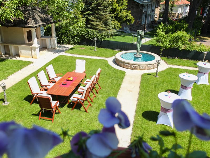 Bašta Muse Event House & Garden sa drvenim stolom, kokteil stolovima, fontanom i skulpturom