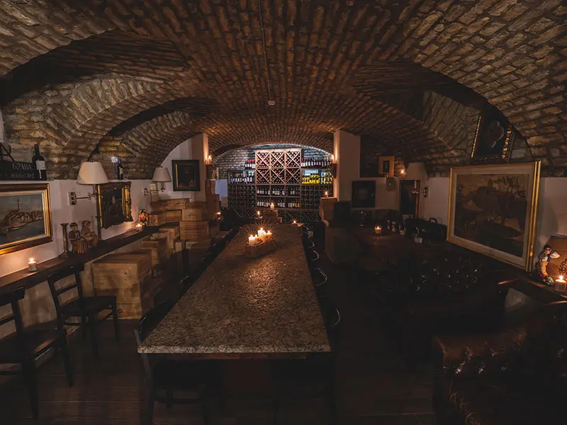 Vinski podrum u Artist Hotel Subotica sa ciglenim svodovima, dugačkim granitnim stolom, svećama, umetničkim slikama i toplim ambijentalnim osvetljenjem