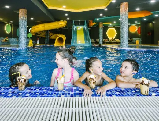 Deca sa milkšejkovima u unutrašnjem bazenu aqua parka hotela Kraljevi Čardaci Spa sa žutim i tirkiznim toboganima u pozadini