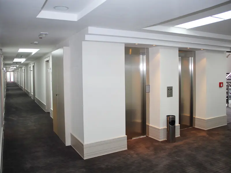 Hodnik sa liftovima u Hotel Vojvodina sa belim zidovima, sivim tepihom, čeličnim vratima liftova i modernim plafonskim osvetljenjem