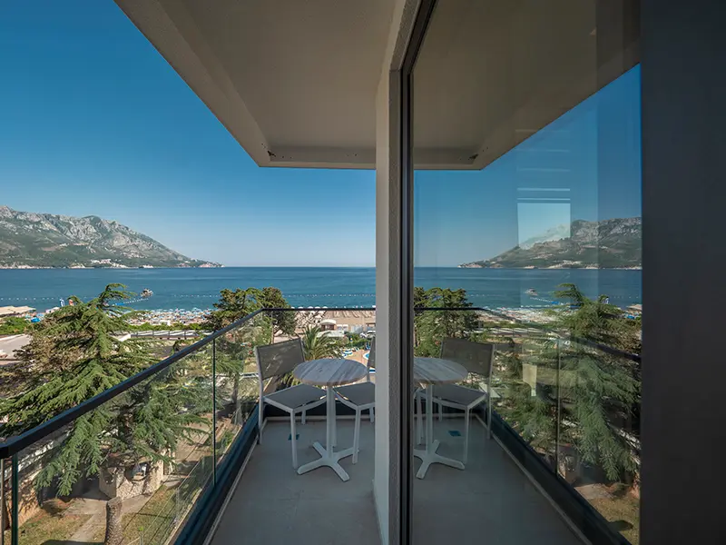 Balkon sobe u Hotel Montenegro sa staklenom ogradom, belim stolom i stolicama i panoramskim pogledom na more, plažu i planine Bečića