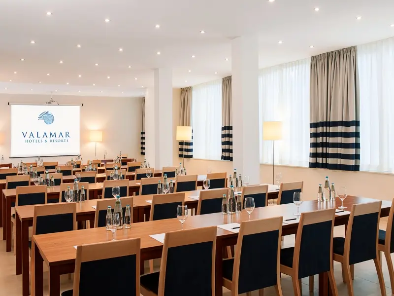 Konferencijska sala u Valamar Sanfior Hotel u učioničkom rasporedu sa drvenim stolovima, tamnoplavim stolicama, projektorom sa Valamar logom i prugastim zavesama