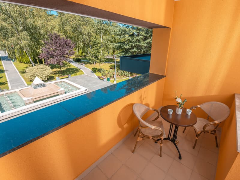 Balkon sobe u Garden City Hotel Konjic & Resort sa narandžastim zidom, stolićem, stolicama i pogledom na fontanu i zeleni park