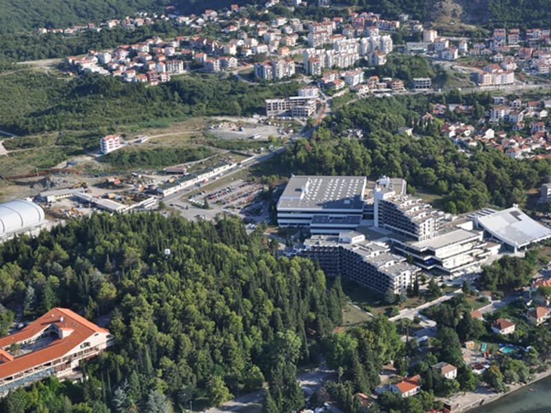 Panoramski pogled iz vazduha na Institut Dr Simo Milošević A.D. Igalo sa kompleksom zgrada okruženim zelenilom, brdom i okolnim naseljima