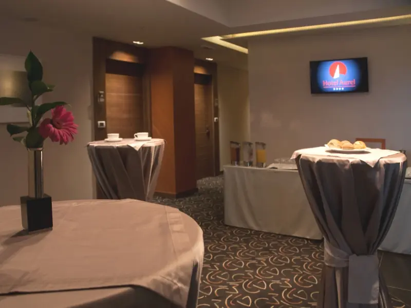 Koktel sala u Hotel Aurel sa visokim koktejl stolovima, TV ekranom sa logom hotela, pecivima i ružičastim cvetom