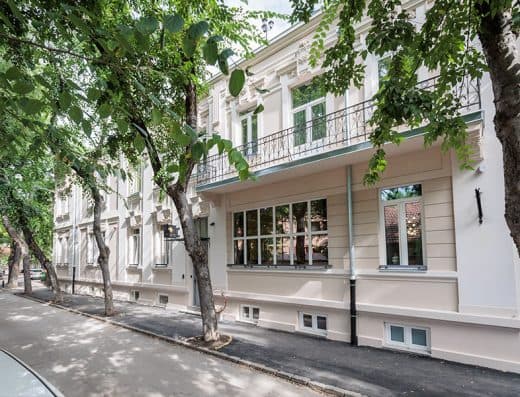 Spoljašnjost Artist Hotel Subotica sa belom klasičnom fasadom, balkonom sa kovanom ogradom i drvoredima na ulici