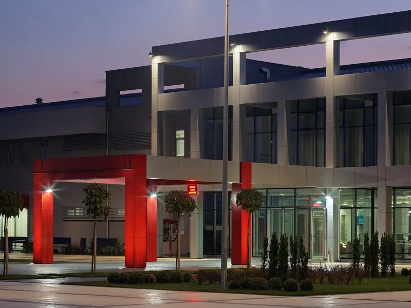 Eksterijer Hotel Srbija Lux sa modernom sivom fasadom, crvenim ulaznim portalom, dekorativnim drvećem i noćnim ambijentalnim osvetljenjem