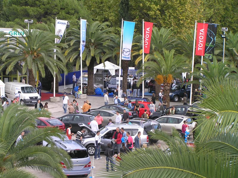 Auto izložba na otvorenom prostoru Jadranski sajam sa vozilima, zastavama brendova, posetiocima i palmama