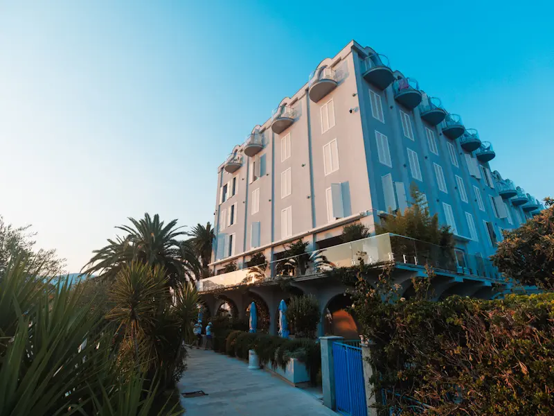 Eksterijer Hotel Palma danju sa fasadom u svetlim tonovima, balkonima, lučnim prolazima, palmama i zelenilom