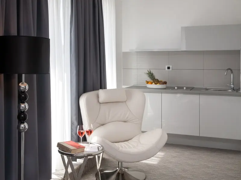 Apartman u Hotel Aurel sa belom kožnom foteljom, čašama crnog vina, kuhinjskim elementima i dizajnerskom stonom lampom