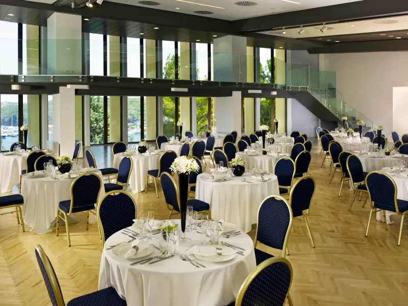 Banket sala u Hotel Park Plaza Histria Pula sa okruglim stolovima, plavim stolicama, belim stolnjacima, cvećem i galerijom na spratu