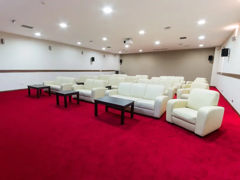 Bioskopska sala u Garden City Hotel Konjic & Resort sa belim kožnim kaučima i foteljama, crnim stolićima, crvenim tepihom i zvučnicima na zidu