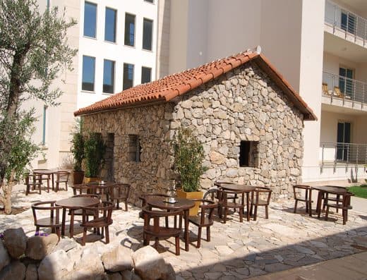 Spoljni pogled na kamenu etno kuću u Hotel Splendid Conference & Spa Resort sa crepovima, drvenim stolovima i stolicama na terasi i stablom masline