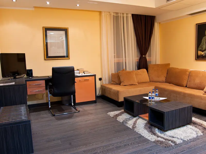 Dnevna soba apartmana u Hill Hotel sa narandžastim kaučem, crnom kožnom radnom stolicom, TV-om, drvenim podom i portretom na zidu