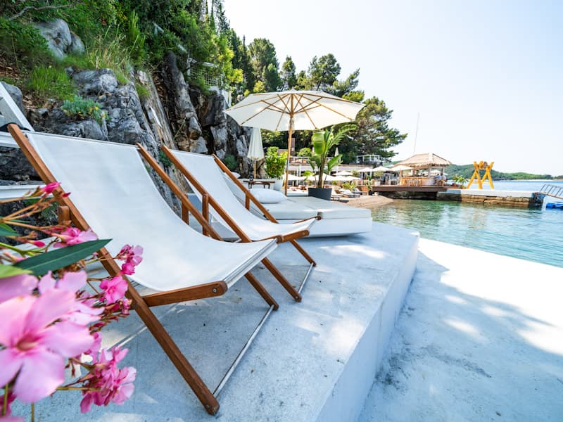 Drvene deck stolice sa belim jastucima na obali Escape Plaža i Restoran sa ružičastim cvećem, suncobranima i pogledom na more