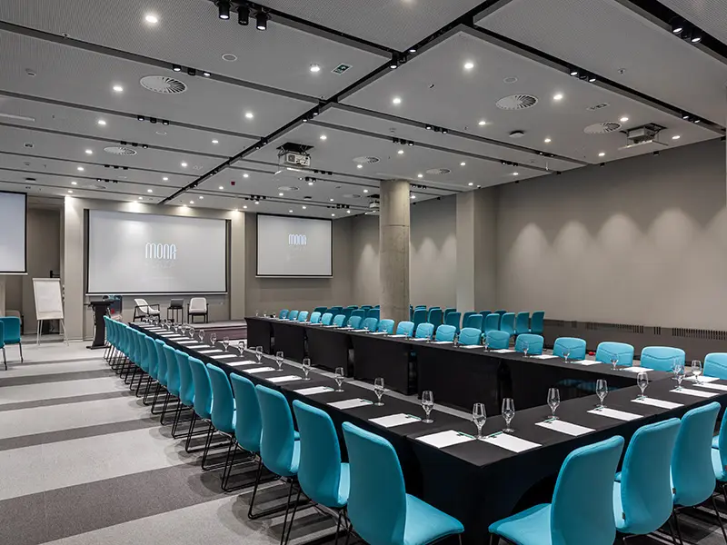 Konferencijska sala u Hotel Mona Plaza sa tirkiznim stolicama u učioničkom rasporedu i Mona Plaza logom na dva ekrana