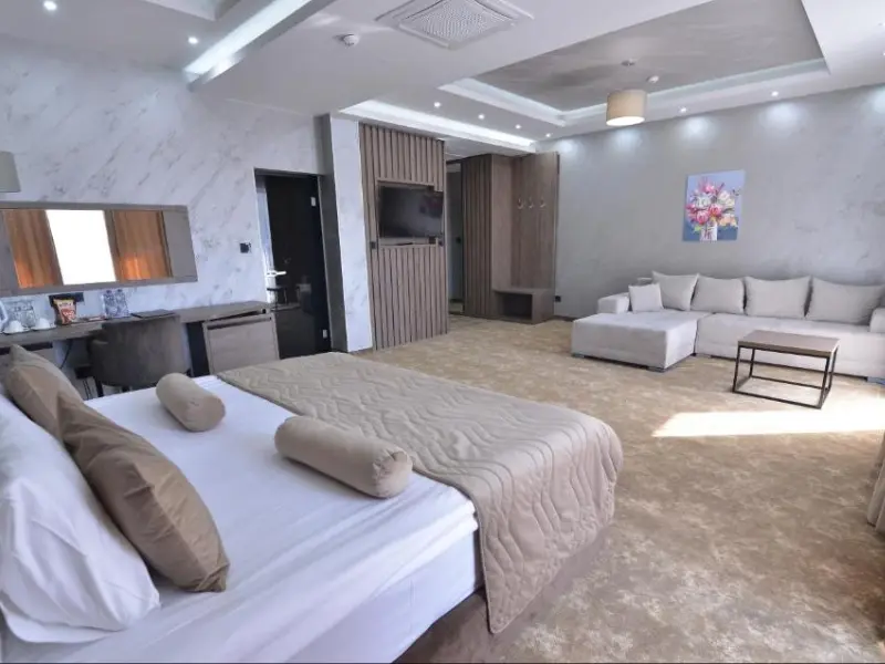 Prostrani apartman u Hotel Vrnjačke Terme sa bračnim krevetom, bež prekrivačem, sivim ugaonim kaučom, TV-om, mermernim zidovima i slikom cveća