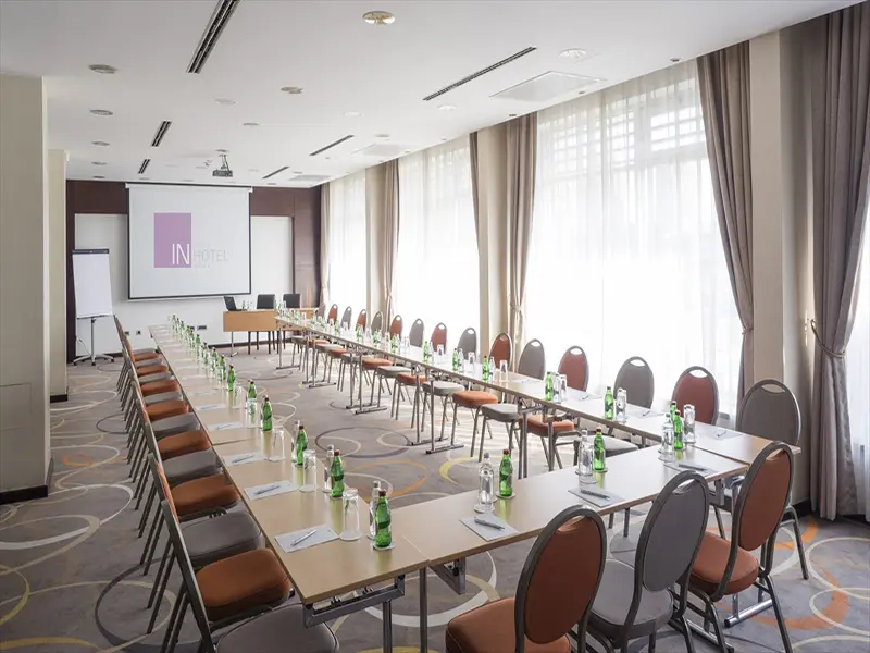Konferencijska sala u IN Hotel sa stolovima u U-rasporedu, projektorskim platnom sa logom hotela, velikim prozorima i vodom na stolovima