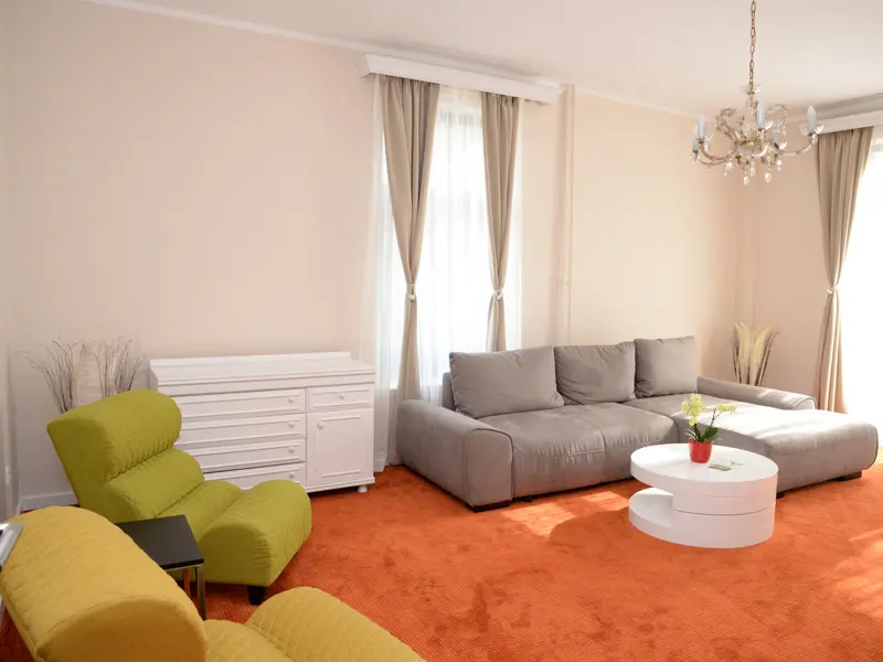 Dnevna soba apartmana u Zepter Hotel Vrnjačka Banja sa sivim kaučem, zelenom foteljom, narandžastim tepihom, belom komodom i kristalnim lusterom