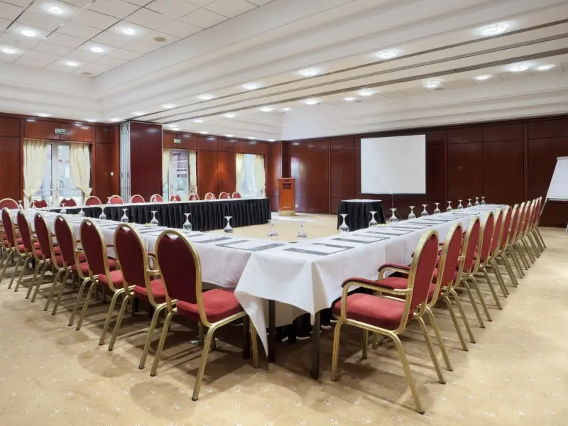 Konferencijska sala u Crowne Plaza Bucharest Hotel sa crvenim stolicama u U-rasporedu, belim stolnjacima, drvenim zidnim panelima, projekcionim platnom i govornicom