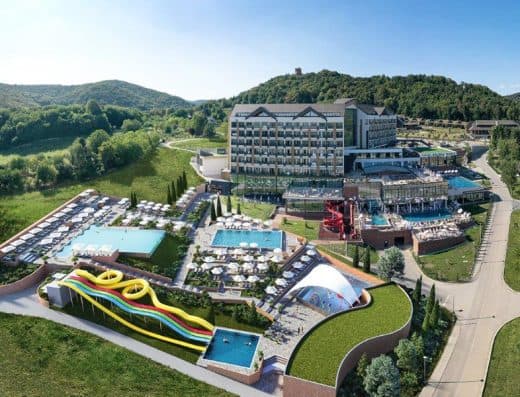 Pogled iz vazduha na ceo kompleks Mövenpick Resort & Spa Fruške Terme sa spoljnim bazenima, toboganima, hotelskom zgradom i zelenim brdima Fruške gore