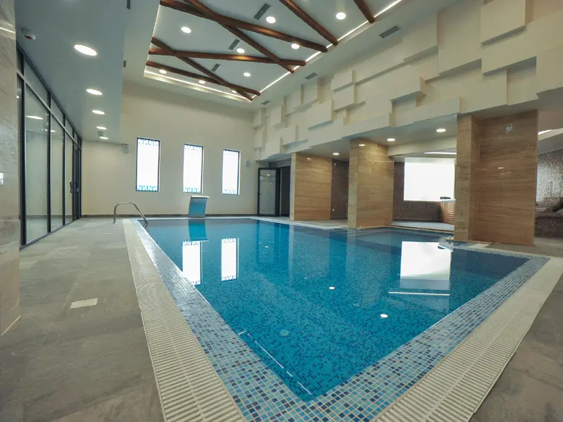 Unutrašnji bazen u Hotel Nais sa plavim mozaikom, belim geometrijskim zidnim panelima, drvenim stubovima, visokim plafonom i prozorima