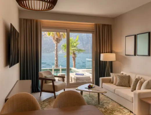 Dnevna soba apartmana u Hyatt Regency Kotor Bay Resort sa bež kaučem, drvenim stolom, foteljom i pogledom na Boku Kotorsku kroz balkonska vrata