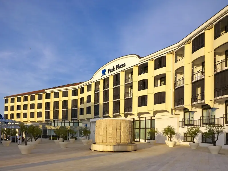 Eksterijer Hotel Park Plaza Histria Pula sa žutom fasadom, natpisom Park Plaza, kamenom skulpturom, maslinovim stablima i trgom ispred