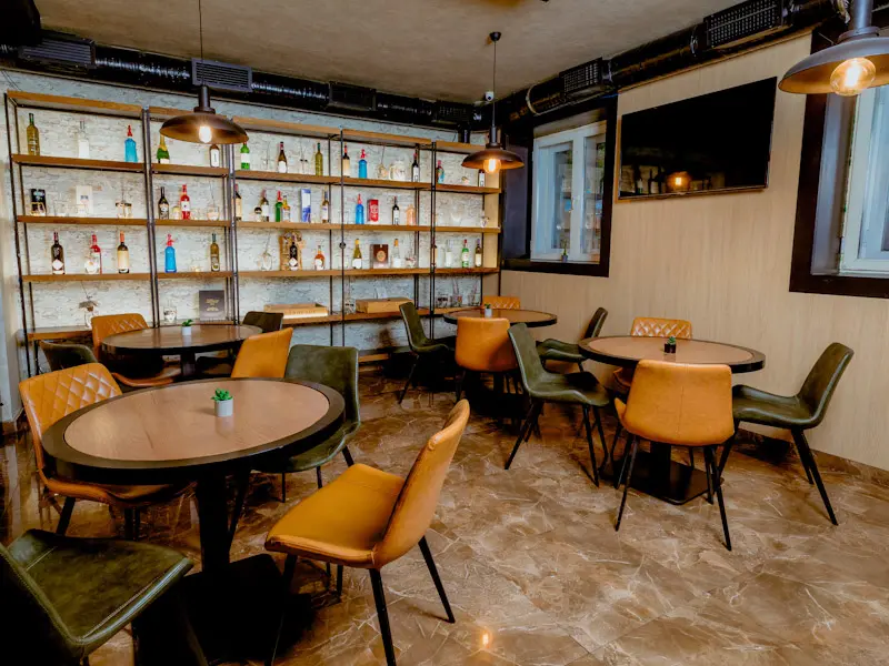 Bar u Hotel Nobel Gallery sa industrijskim stilom, metalnim policama sa flašama pića, okruglim stolovima, kožnim stolicama u oker i zelenoj boji i visećim lampama