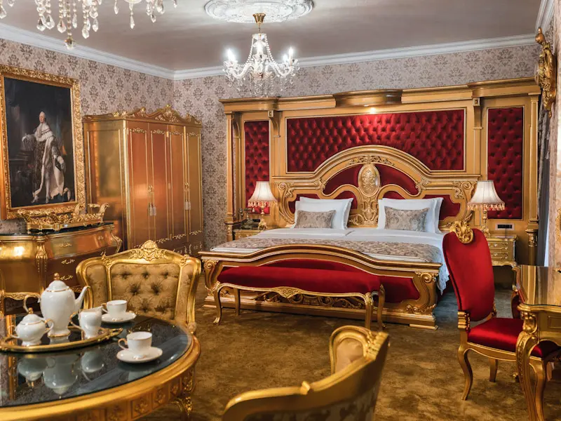 Luksuzni apartman u Prezident Palace Belgrade sa velikim crvenim tapaciranim uzglavljem, zlatnim baroknim nameštajem, kristalnim lusterom, zlatnim ormanom i umetničkom slikom