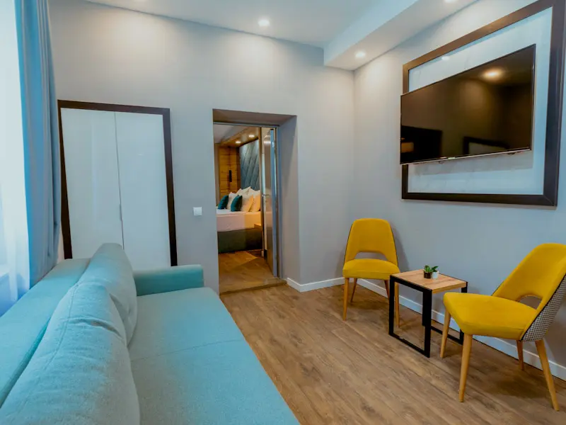Dnevna soba apartmana u Hotel Nobel Gallery sa svetloplavom sofom, žutim stolicama, TV-om na zidu, drvenim podom i pogledom na spavaću sobu
