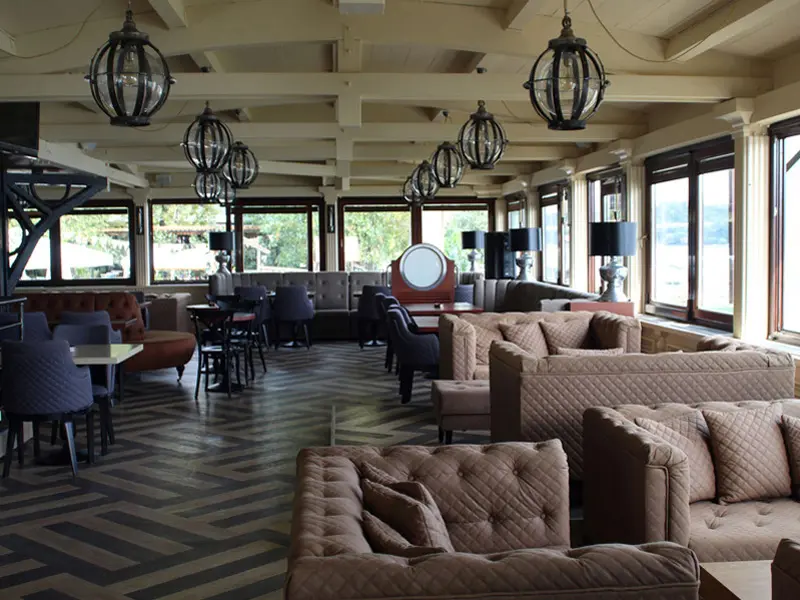 Lobi bar u Turističko naselje Ribarsko ostrvo sa kapetanskim sofama, okruglim lampama, drvenim podom i pogledom kroz prozore