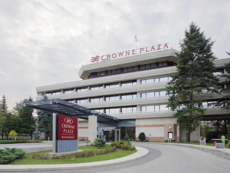 Eksterijer Crowne Plaza Bucharest Hotel sa svetlećim natpisom na krovu, crvenim info tablom na ulazu, kružnim prilazom, zelenim travnjakom i drvoredima