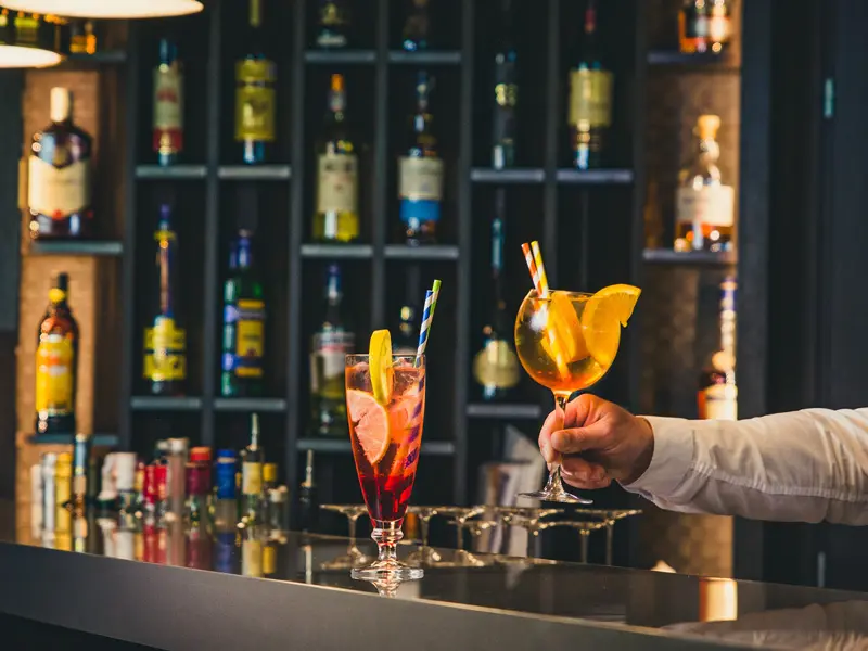 Barmen servira koktele u baru Hotel voco Podgorica sa šarenim koktelima, policama sa flašama pića i toplim ambijentalnim osvetljenjem