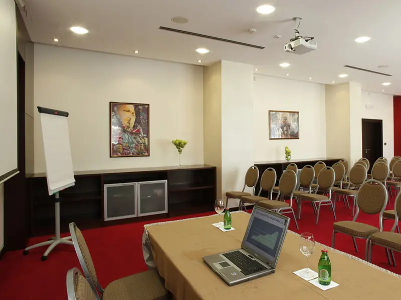Konferencijska sala u IN Hotel sa projektorom, flipchartom, crvenim tepihom, stolicama u teatar rasporedu i umetničkim slikama na zidovima