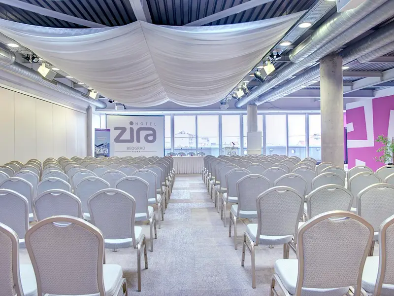 Penthouse sala u Hotel Zira Beograd sa bež stolicama u pozorišnom rasporedu, belim draperima na plafonu, pink dekorativnim zidom, logom hotela i industrijskim osvetljenjem