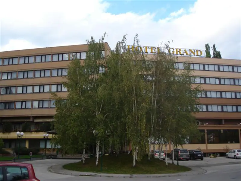 Fasada Hotel Grand Sarajevo sa zlatnim natpisom, brezama ispred zgrade, parkingom i oblačnim nebom