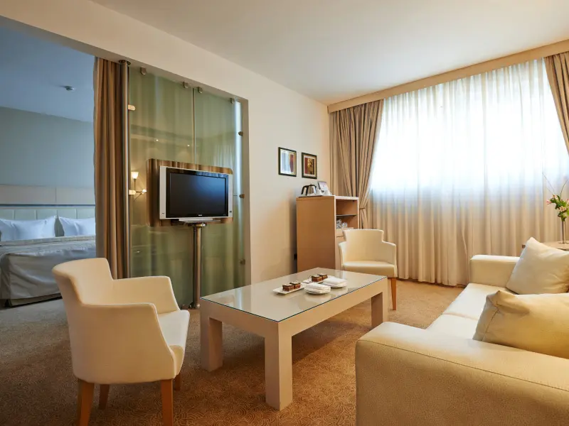 Dnevni boravak apartmana u IN Hotel sa belim foteljama, staklenim stolićem, TV-om na staklenom paravanu i pogledom na spavaću sobu