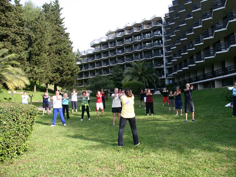 Grupna gimnastika na travnjaku ispred Institut Dr Simo Milošević A.D. Igalo sa instruktorom, gostima i pogledom na fasadu zgrade sa balkonima