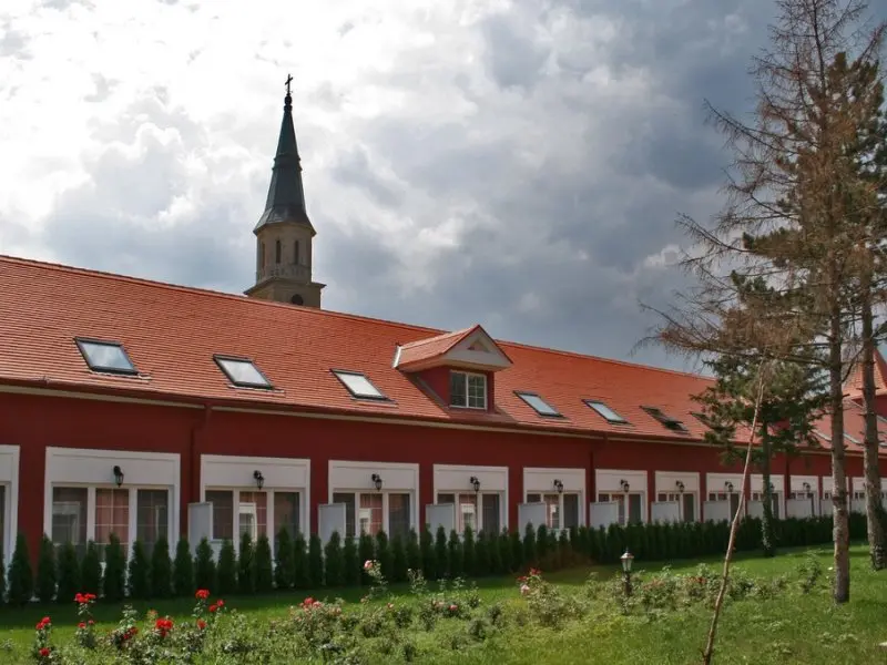 Bočni pogled na Lovački Dvorac Kaštel Ečka sa crvenom zgradom, narandžastim krovom, krovnim prozorima, zvonikom crkve i zelenilom