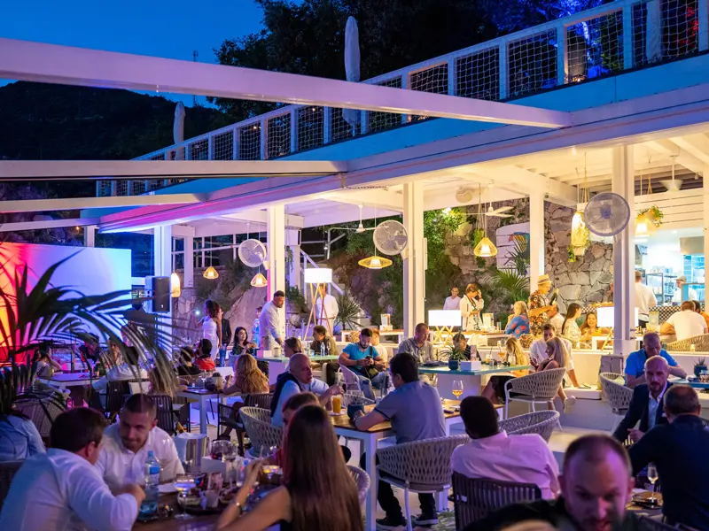 Noćni ambijent restorana Key West Beach & Restaurant sa plavim osvetljenjem, belim nameštajem i gostima